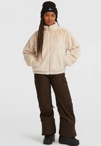 Beige, textuurvolle fleecejas met hoge kraag en ritssluiting aan de voorkant, gecombineerd met bruine cargobroek en zwarte laarzen. Zwarte beanie zorgt voor contrast.