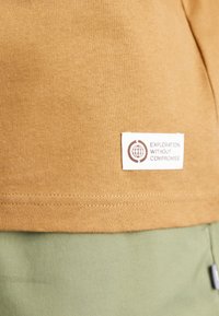 Koszula z krótkim rękawem o bawełnianej fakturze w kolorze musztardowym z zielonym obszyciem. Posiada białą metkę z napisem "Eksploracja bez kompromisów".