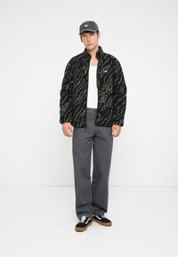 Black and green patterned fuzzy jacket with a zip, white t-shirt, gray trousers, and black sneakers with white laces and tan soles.