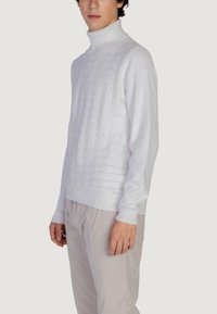 Bijela pletena turtleneck vesta s uzorkom, uskog kroja i rebrastim manžetama, uparena sa svijetlim prugastim hlačama.