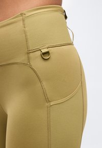 Κοντινό πλάνο ατόμου που φοράει ψηλόμεσα κολάν μουσταρδί με πλαϊνή τσέπη και μικρή λεπτομέρεια D-ring κοντά στη μέση.