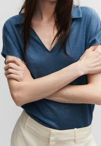 Tyrkysové tričko z lněné směsi s výstřihem do V a krátkým rukávem, které má volnější střih a měkkou texturu, v kombinaci se světlými kalhotami.