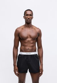 Črne bokserice z belim elastičnim pasom, na katerem je napis "Calvin Klein". Model ima mišičasto postavo in nevtralen izraz obraza.