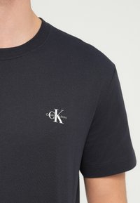 Tmavomodré tričko s malým bielym logom "cK Calvin Klein Jeans" potlačeným na ľavej strane hrude, ktoré nosí osoba so svetlou pokožkou.