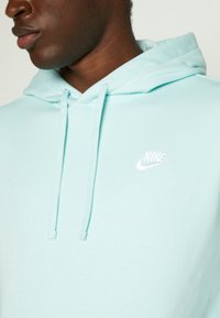Primer plano de una persona que lleva una sudadera con capucha Nike de color azul claro con el logo blanco en el pecho y cordones colgando de la capucha.