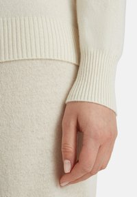Maglione di colore crema lavorato a maglia, con polsini a coste e una finitura morbida e testurizzata. Primo piano di una mano appoggiata sul tessuto.