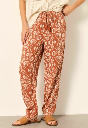 Rust-colored loose pants with white floral patterns, elastic waistband with tassel ties, worn with woven slide sandals and a cream blouse.
