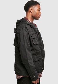 Black hooded jacket featuring a button closure, front pockets, and a textured fabric. Layered over a plaid shirt, viewed from the side.