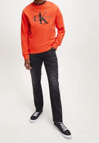 Pomarańczowa bluza z czarnym logo Calvin Klein Jeans, zestawiona z ciemnymi dżinsami i czarnymi trampkami z białymi sznurowadłami oraz fakturowanym wzorem.