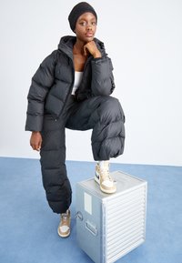 Svart puffjacka och byxor med fyrkantig form, quiltad textur. Vit topp och ljusbruna högskaftade sneakers. Grå metallåda under.