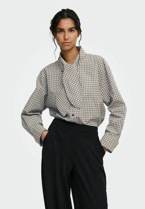 Femme aux cheveux foncés portant un chemisier à manches longues à carreaux avec un grand nœud et un pantalon noir taille haute, debout les mains dans les poches.