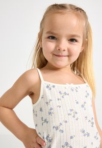 Valkoinen hihaton paita, jossa on sininen kukkakuosi, spagettiolkaimet ja häilyvä nauhakoriste. Kevyt kangas vaakasuoralla raidalla.