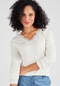 Licht romige, lange mouw top met een V-hals en fijne kantafwerking. Zachte stof, getailleerde stijl, subtiele textuur, gecombineerd met blauwe jeans.