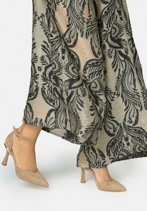 Pies con tacones de ante beige con pulsera en el tobillo, combinados con una falda fluida que presenta un patrón de flores y hojas negras sobre fondo beige.