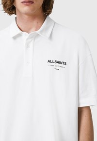 Hvit polo skjorte laget av bomull, med klassisk krage, tre-knapps placket og svart "ALLSAINTS"-logo med japansk tekst.