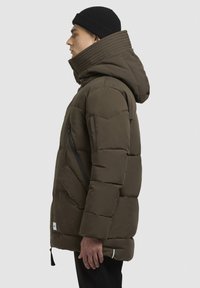 En pufferjakke i mørke oliven grøn med quiltet design, stor hætte, lynlås-lommer og et label ved sømmen. Tekstureret stoffinish.