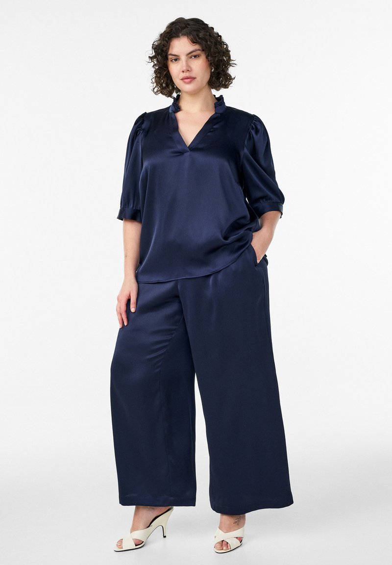 Γυναίκα στέκεται φορώντας παντελόνι από νάιλον σε σκούρο μπλε με φαρδιά μπατζάκια και ταιριαστή μπλούζα με βολάν στο λαιμό, καθώς και λευκά σανδάλια με τακούνια.