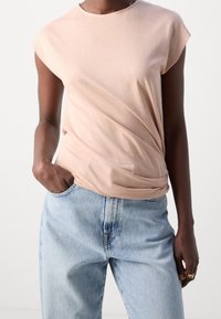 Camiseta de algodón rosa claro con mangas cortas y corte relajado, que presenta un detalle torcido en la cintura, combinada con unos jeans azul claro.