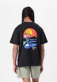 Sort bomulds T-shirt med et farverigt grafisk print af en solnedgang, bjerge og et cocktailglas på bagsiden. Korte ærmer, afslappet pasform.