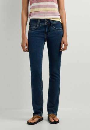 Frau trägt dunkelblaue Jeans kombiniert mit braunen Sandalen und einem ärmellosen gestreiften Oberteil, steht vor einem schlichten Hintergrund.