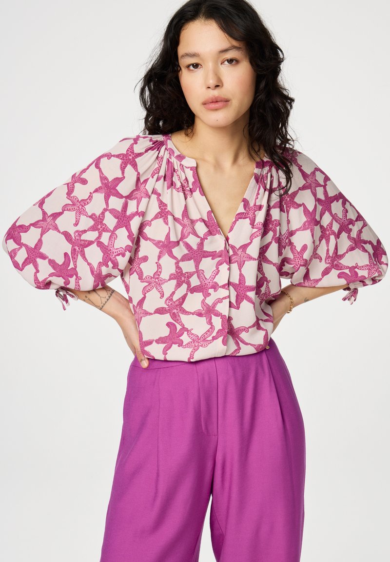 Roze blouse met een zeesterpatroon, V-hals en korte pofmouwen. Gecombineerd met paarse broek met hoge taille. Soepele stoftextuur.