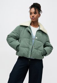 Casaco acolchoado verde de bombazina com gola de pêlo falso creme, textura acolchoada horizontal, botões de pressão prateados e bolsos frontais.