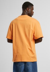Orange bomulds T-shirt med rund halsudskæring og korte ærmer, vist fra ryggen, parret med blå denimshorts.