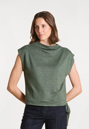 Femme portant un haut vert sans manches noué sur le côté et un pantalon noir, debout avec les mains derrière le dos, regardant légèrement sur le côté.