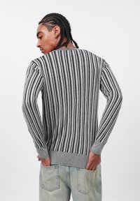Uomo con capelli intrecciati che indossa un maglione a righe verticali grigio e nero e jeans azzurri chiari, con le mani nelle tasche posteriori, di spalle.