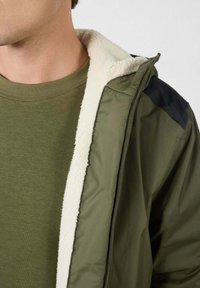 Olijfgroene jas met zwarte accenten op de schouders, voorzien van een crèmegekleurde fleece voering en een ritssluiting. Draagt over een bijpassend olijfgroen T-shirt.