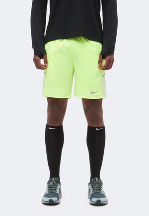 Sportowiec noszący neonowo żółte spodenki sportowe, czarną bluzkę z długim rękawem, czarne skarpety Nike do kolan oraz szare i błękitnozielone buty do biegania, stojący.