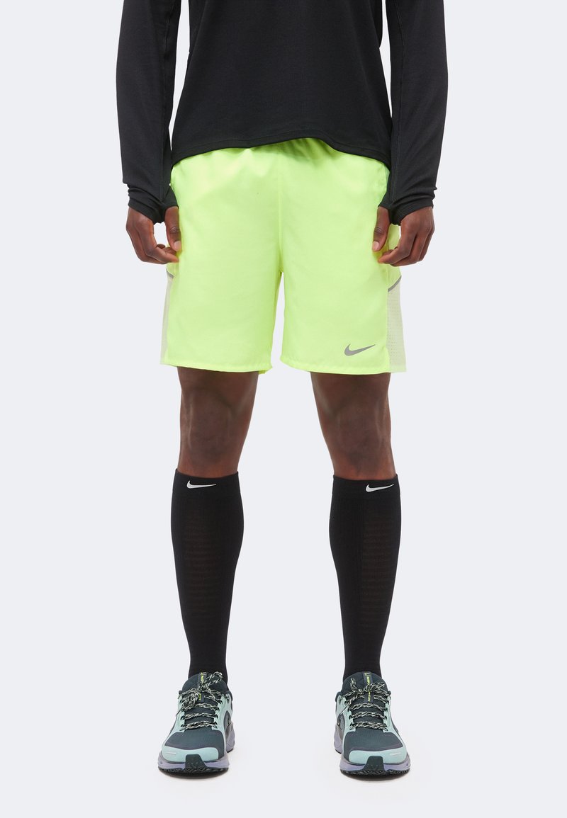 Athlète portant un short de sport jaune fluo, un haut à manches longues noir, des chaussettes Nike noires montantes aux genoux et des chaussures de course grises et bleu sarcelle, debout.