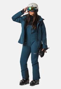 Skioutfit in Dunkeltürkis, bestehend aus einem figurbetonten Schneesuit mit Reißverschlüssen, einer schwarzen Basisschicht, Helm und bunten Skibrillen. Enthält Skischuhe.