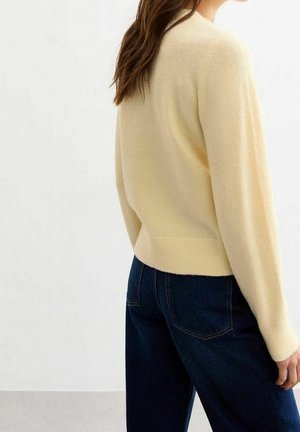 Femme aux longs cheveux bruns portant un pull crème et un jean bleu foncé, se tenant devant un fond clair uni.
