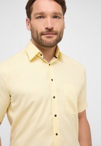 Camicia a maniche corte di colore giallo chiaro con un motivo a texture, dotata di tasca sul petto e bottoni neri lungo il davanti.