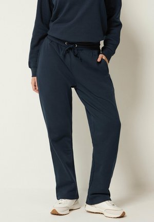 Persoon met navyblauwe joggingbroek met trekkoord, bijpassende sweatshirt en witte sneakers, staand tegen een effen lichte achtergrond.