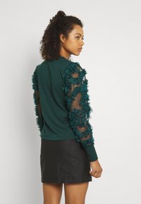 Green sweater with sheer, textured sleeves decorated with fabric flowers, worn with a black skirt. Features a round neckline and fitted cuffs.