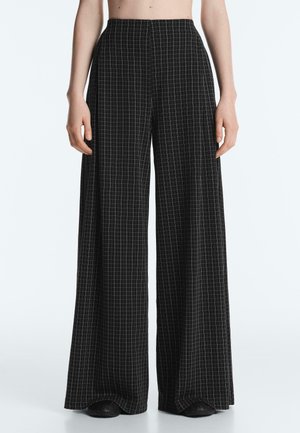 Pantaloni neri a gamba larga con motivo a griglia bianca indossati da una persona con il ventre nudo, che si estendono per coprire scarpe nere.