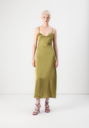 Robe nuisette en satin vert olive avec de fines bretelles, silhouette épurée, coupe longue jusqu'au sol ; associée à des talons hauts à lanières violets.