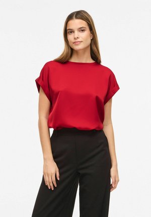 Femme aux cheveux raides portant un chemisier rouge à manches courtes et un pantalon noir taille haute, debout devant un fond blanc uni.