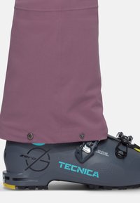 Purple waterproof ski pant covering a grey Tecnica boot with blue and yellow accents, featuring metal snaps and textured detailing.