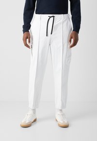 Λευκό παντελόνι cargo με ελαστική μέση και μαύρο κορδόνι, πλαϊνές τσέπες με φερμουάρ και σφιχτές μανσέτες. Φορεμένο με λευκά αθλητικά παπούτσια και κάλτσες μέχρι τον αστράγαλο.