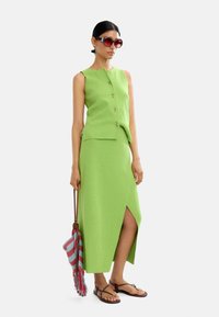 Zielony dzianinowy top bez rękawów z guzikami, pasująca spódnica z bocznym rozcięciem, brązowe sandały i paskowana torba w czerwono-niebieskie paski.