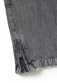 Chaqueta de mezclilla negra con un acabado texturizado, cierre lateral con cremallera y detalles de dobladillo sin terminar, destacando un diseño moderno y casual.