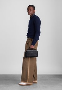 Mężczyzna w granatowym swetrze i szerokich brązowych spodniach trzymający małą czarną torbę, stojący na tle szarego tła.