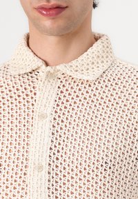 Beige gebreide shirt met een kraag en knoopsluiting, met een open meshpatroon en een zachte, gestructureerde uitstraling.