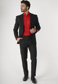 Zwarte pak met een getailleerd ontwerp, gecombineerd met een rode button-up shirt met een subtiel patroon. Zwarte schoenen maken de look compleet.