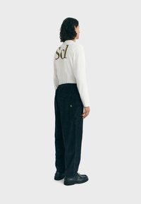 Persona con capelli ricci che indossa una maglietta bianca a maniche lunghe con il testo "Scl" sulla schiena, pantaloni di velluto a coste neri e scarpe nere, in piedi di lato.