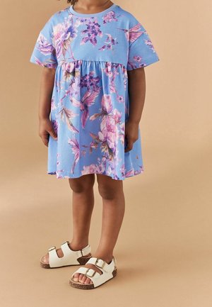 Robe fleurie bleue à manches courtes et taille froncée, associée à des sandales blanches avec deux boucles dorées et semelles en liège.