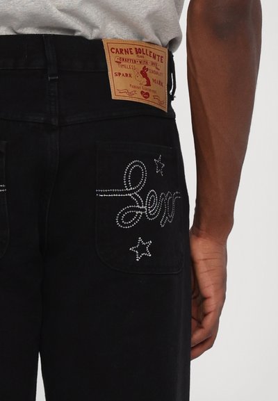 Los jeans de mezclilla negra presentan costuras blancas, con la palabra "sex" y motivos de estrellas en los bolsillos traseros. Una etiqueta color tostado se encuentra en la cinturilla.